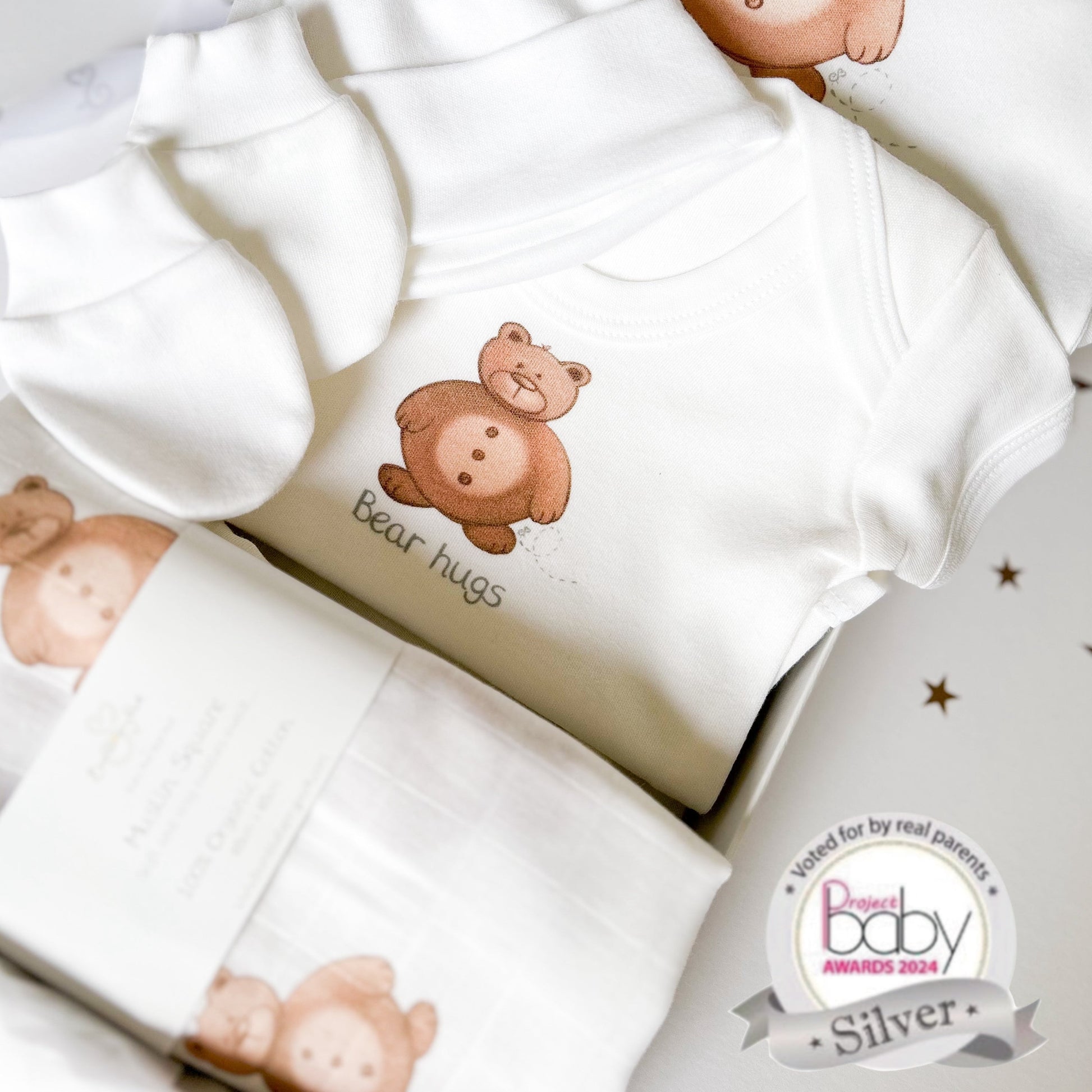 brown teddy bear new baby gift set of a sleepsuit, bodysuit vest, muslin square and hat and mittens with bear hugs slogan, inside a luxury white gift box with satin ribbon gift wrap