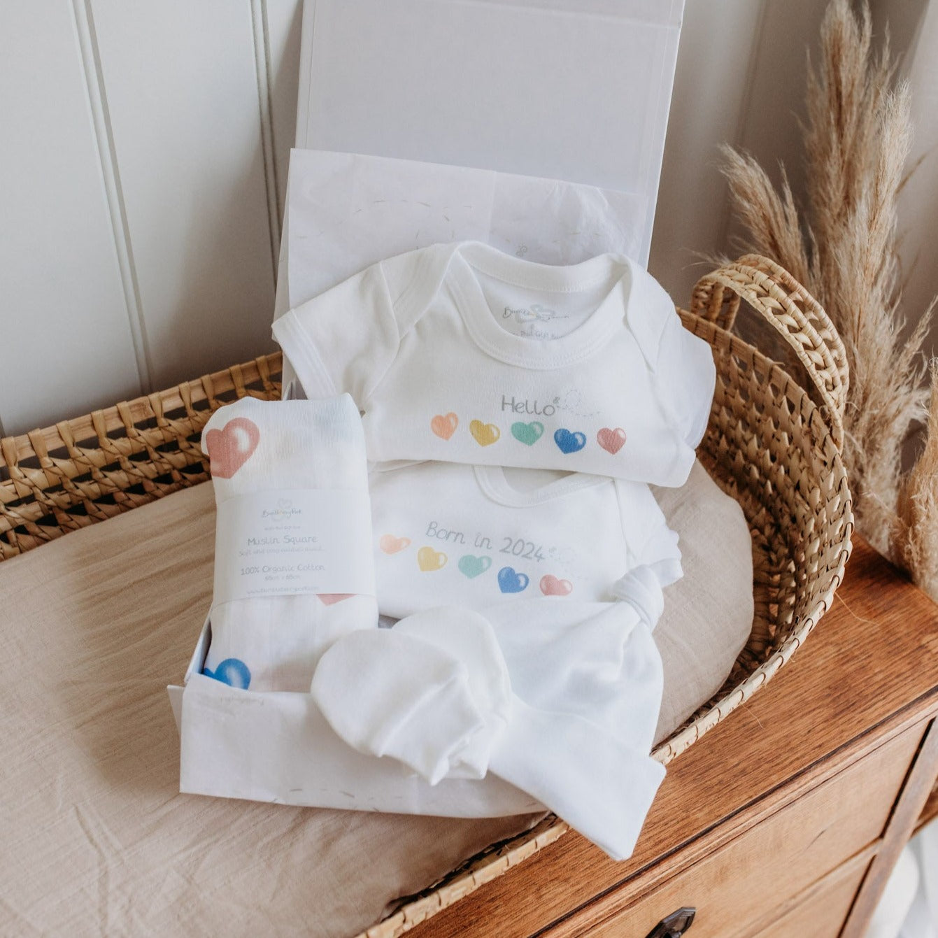 born in 2024 love hearts new baby starter gift set with baby sleepsuit, bodysuit, hat mittens and muslin square with a rainbow colour bright heart design in a luxury white gift box