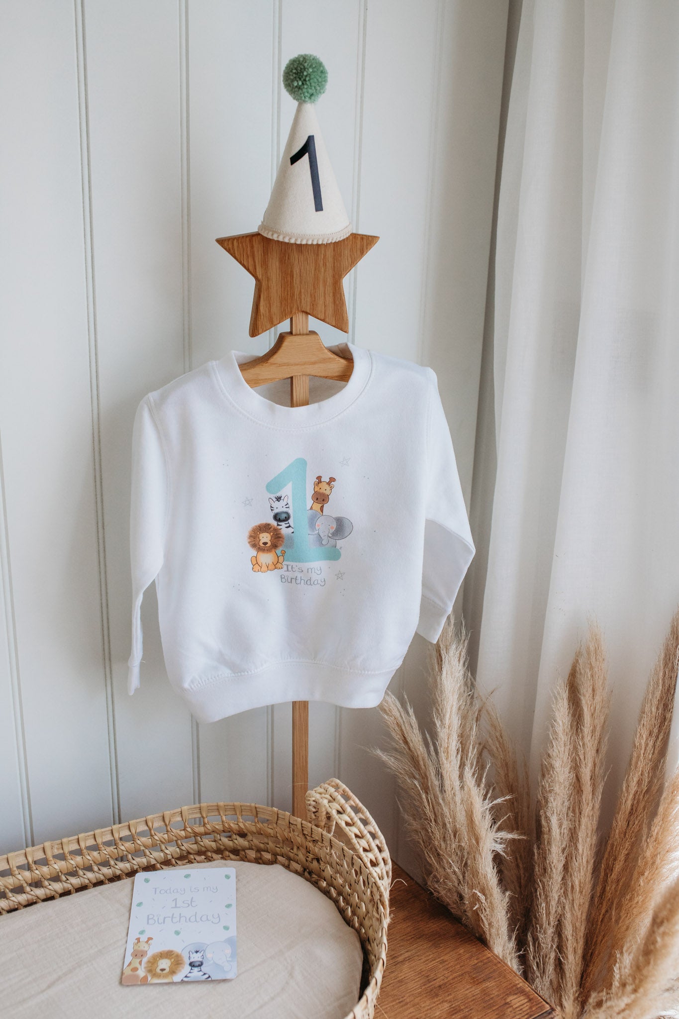 white children's birthday sweatshirt with safari animal 1st birthday design with lion, giraffe, zebra and elephant and neutral colours on a clothes hanger with a 1st birthday party hat