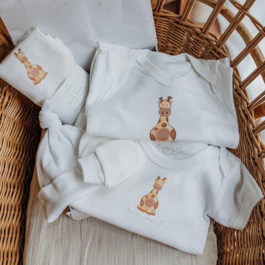 new baby gift set of safari giraffe printed baby clothes including an organic white sleepsuit, vest, hat, mittens and muslin clothes presented inside a baby basket 
