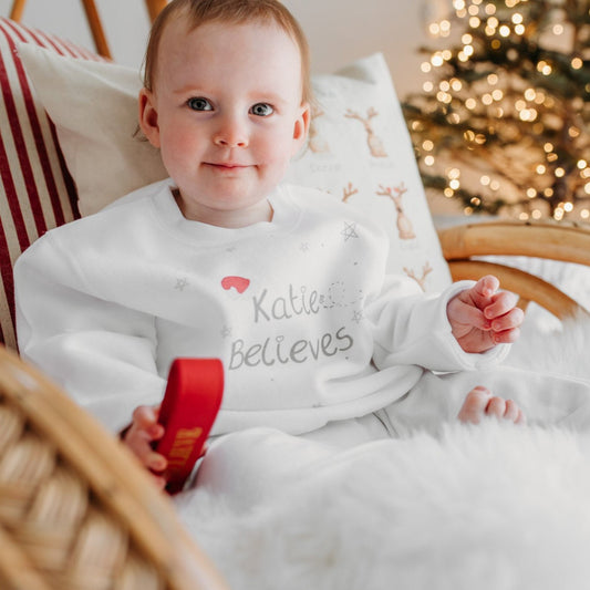 toddler girl wearing a white christmas jumper with "believe" text and a santa's hat on it