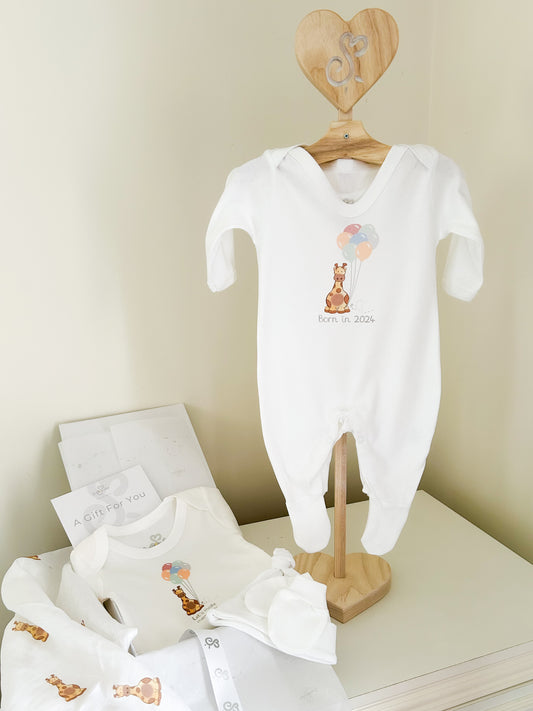 new baby starter gift set including white sleepsuit on a baby clothes stand, a baby vest and muslin square, hat and mittens printed with a safari giraffe and balloons design