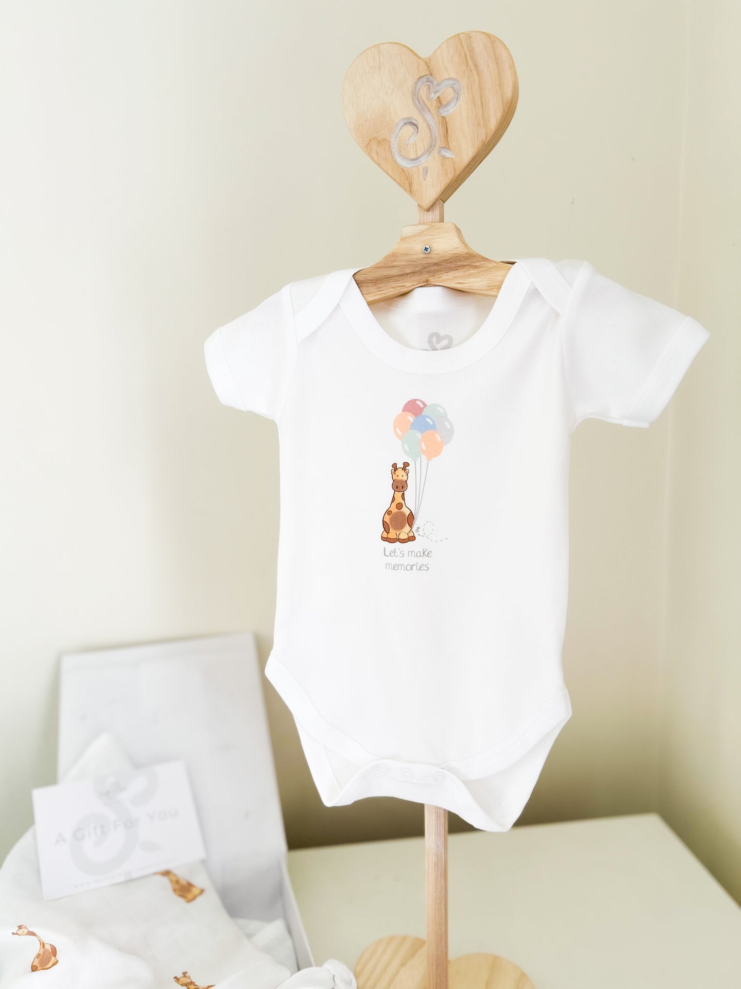children's clothes stand with a white baby vest printed with a cute giraffe images holding some bright colourful balloons and "let's make memories" text