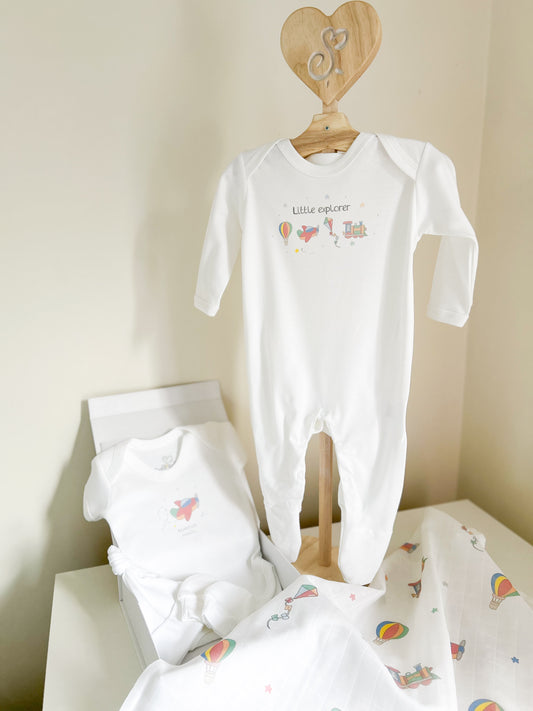 new baby starter gift set including white sleepsuit on a baby clothes stand, a baby vest and muslin square, hat and mittens printed with a little explorers travel and transport and balloons design