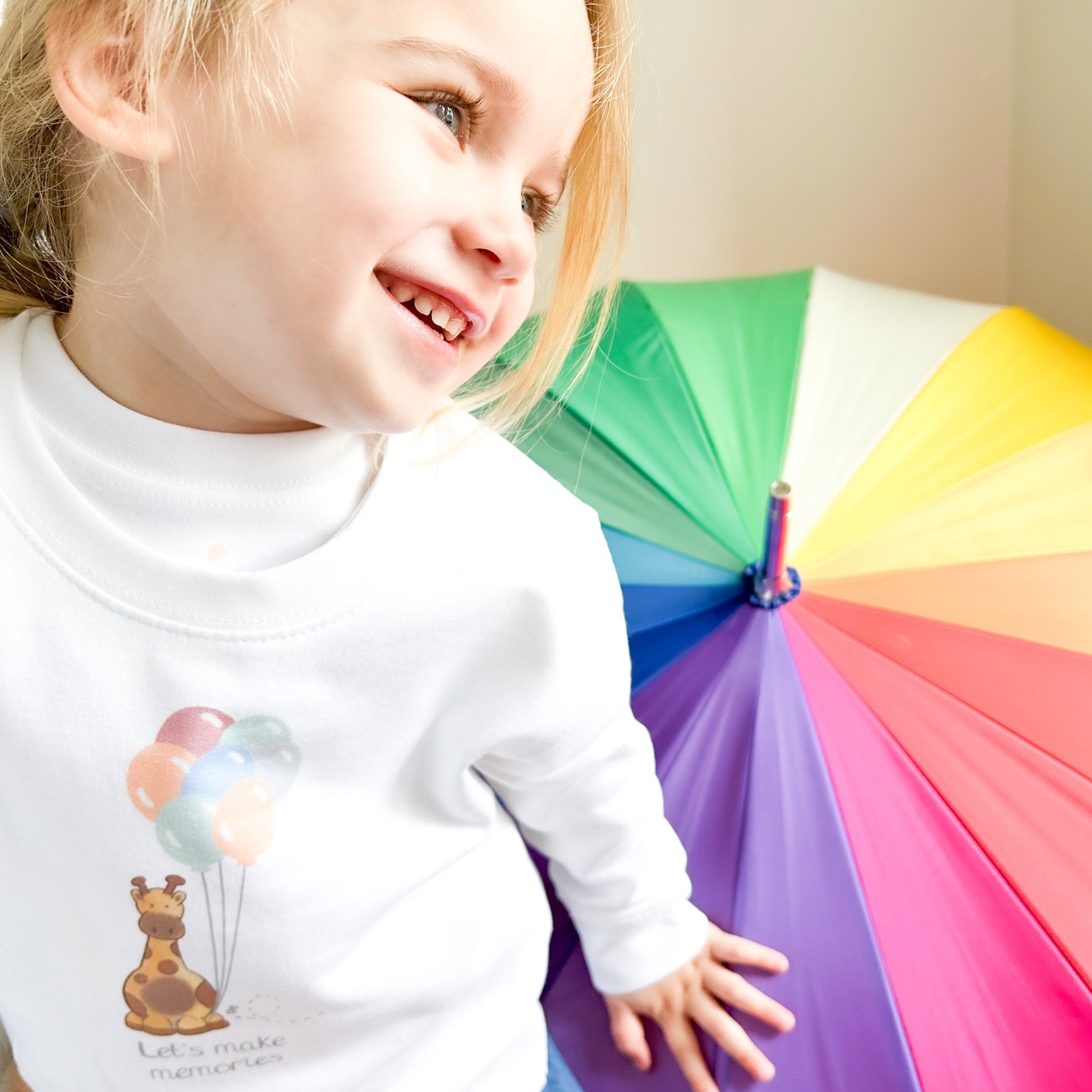 girl wearing a safari giraffe and party balloons sweatshirt smiling next to a rainbow colour umbrella