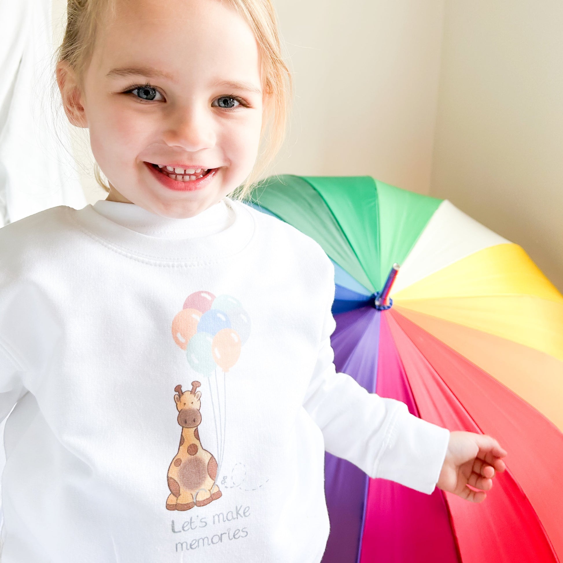 a girl smiling with a colourful umbrella and white sweatshirt on with cute giraffe and balloons design with "let's make memories" quote