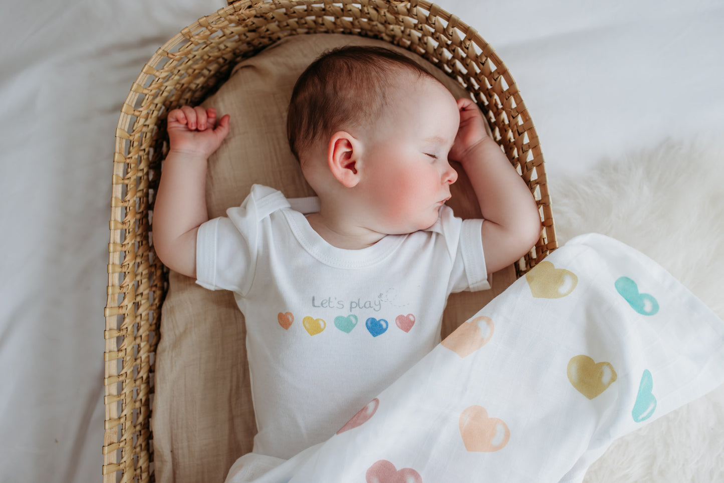 baby asleep lying next to a bright and colourful muslin wrap with bright rainbow coloured love hearts design