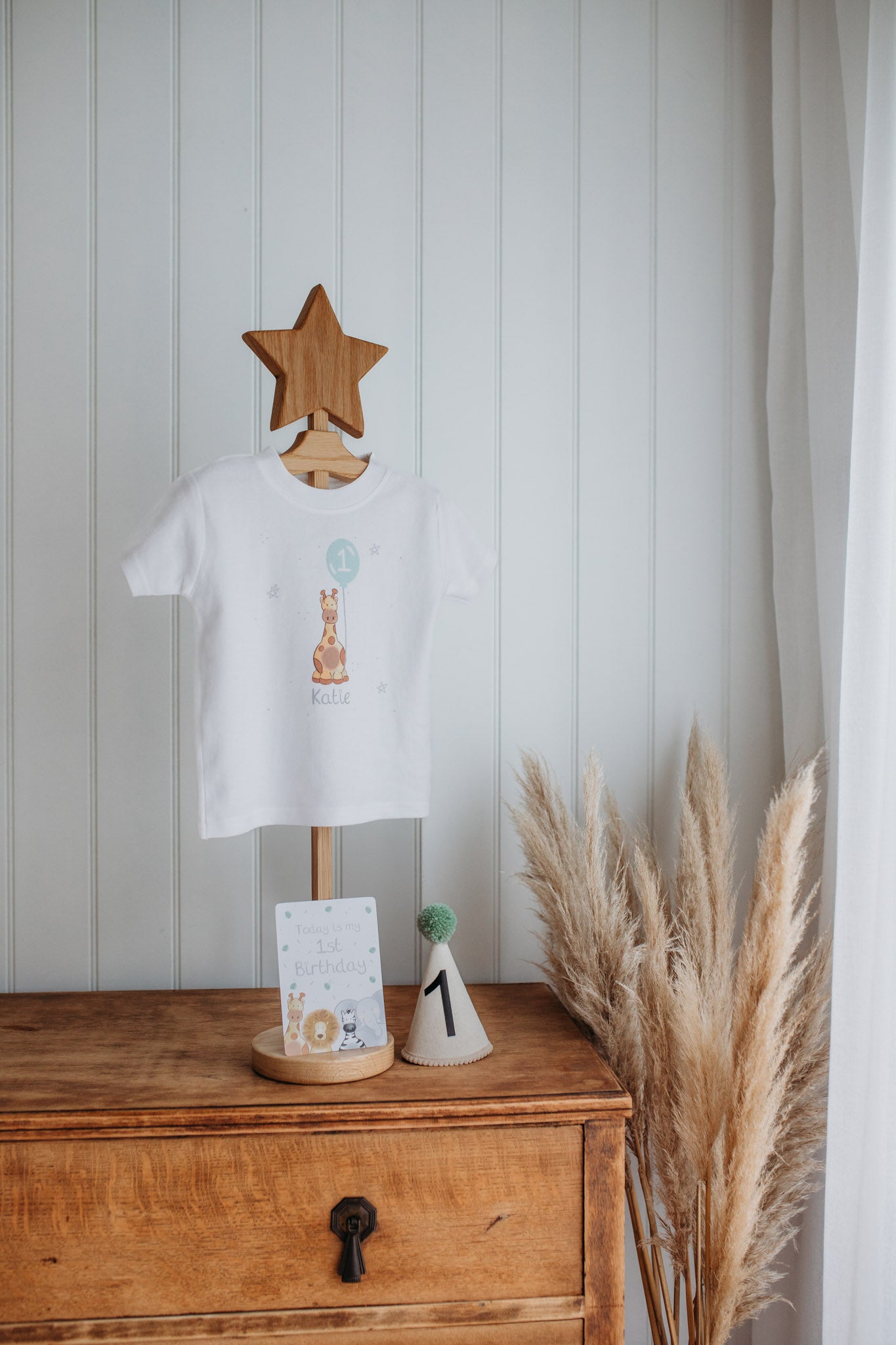 children's wooden clothes stand holding a personalised 1st birthday t-shirt with name katie and safari giraffe printed illustration holding a 1st birthday balloon